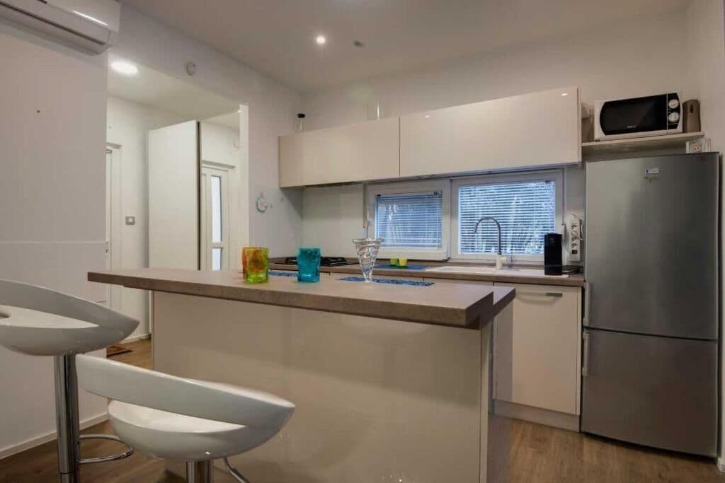 A modern kitchen with white cabinets, a stainless steel refrigerator, a microwave, and a window with blinds.
