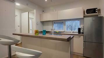 A modern kitchen with white cabinets, a stainless steel refrigerator, a microwave, and a window with blinds.