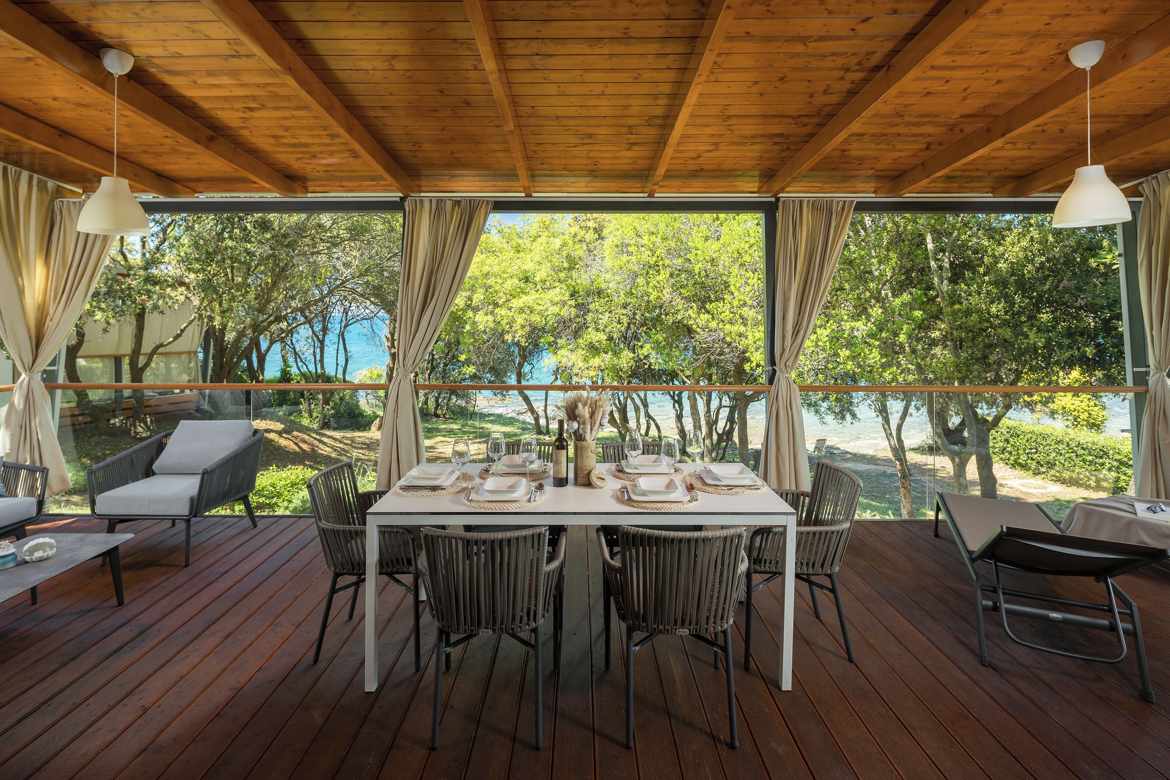 A wooden deck with a table set for dining, surrounded by wicker chairs, overlooking a scenic view of trees and water.