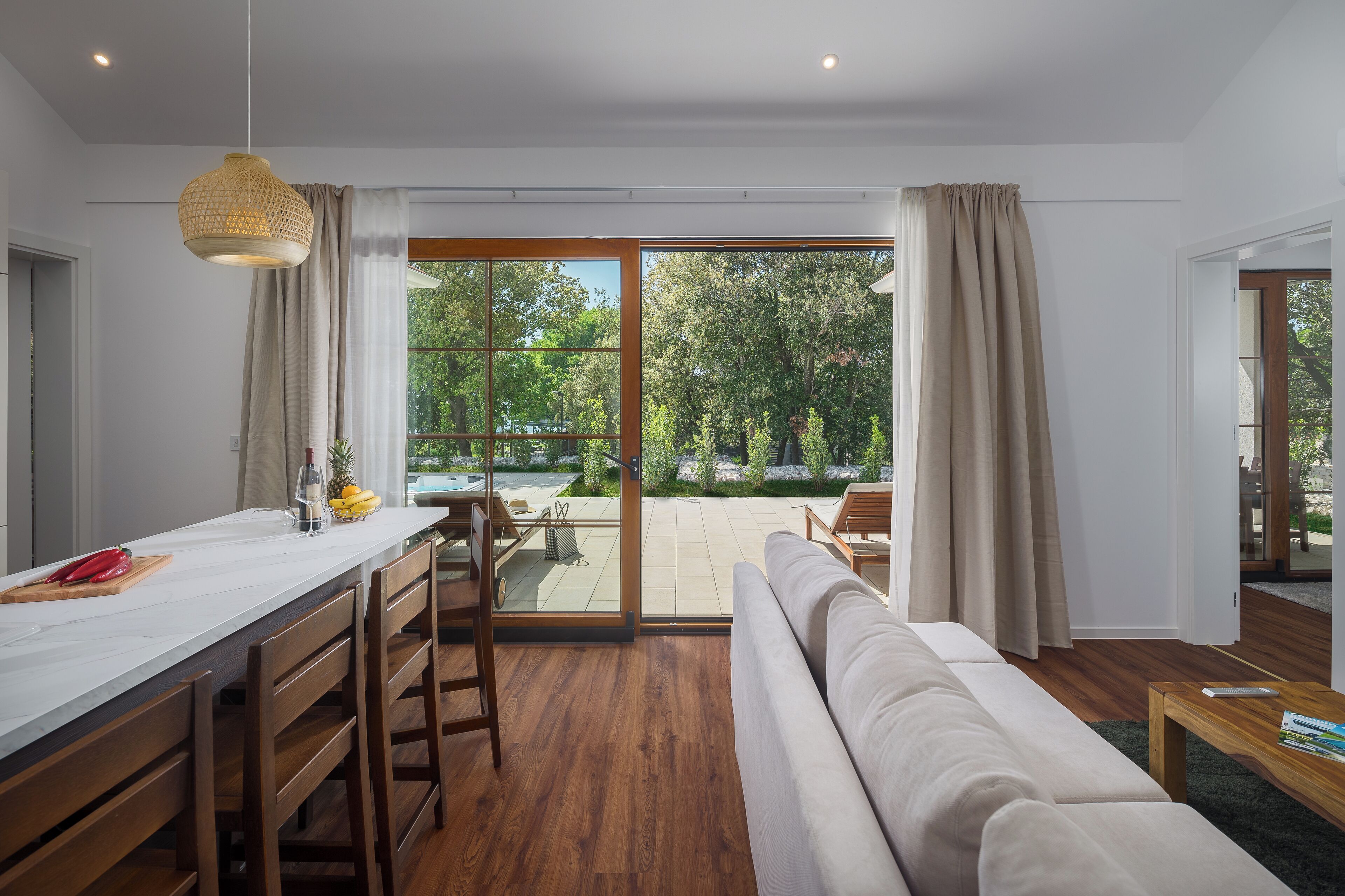 Una sala de estar moderna con un sofá, sillas de madera, una mesa de comedor y un amplio ventanal corredero que da a un espacio exterior.