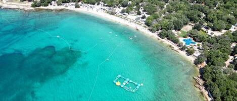 An aerial view of a coastal area with clear turquoise waters, a sandy beach, and dense greenery.