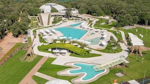 Vista aérea de um resort com várias piscinas, uma estátua de dinossauro e um edifício grande com um design de telhado único.