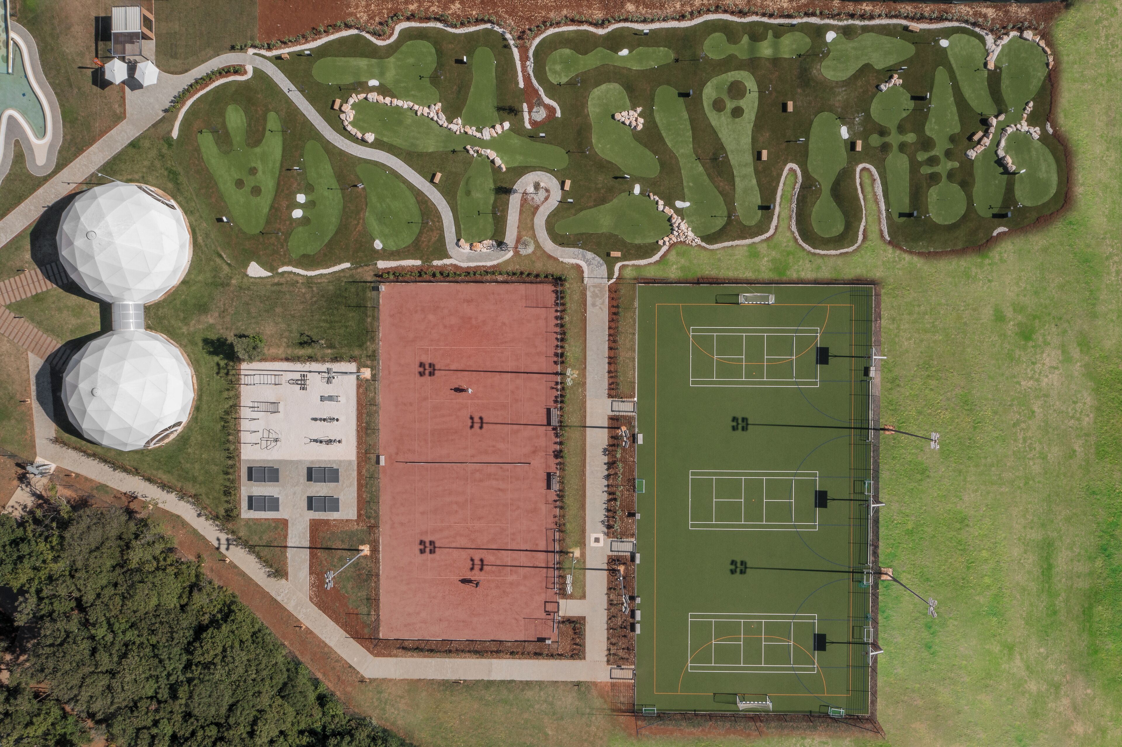 Una vista aerea di un complesso sportivo con un campo da tennis, un campo da basket e un grande edificio con tetto rosso.