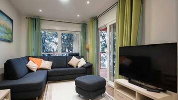 A modern living room with a sectional sofa, ottoman, flat-screen TV, and a sliding glass door to the outdoors.