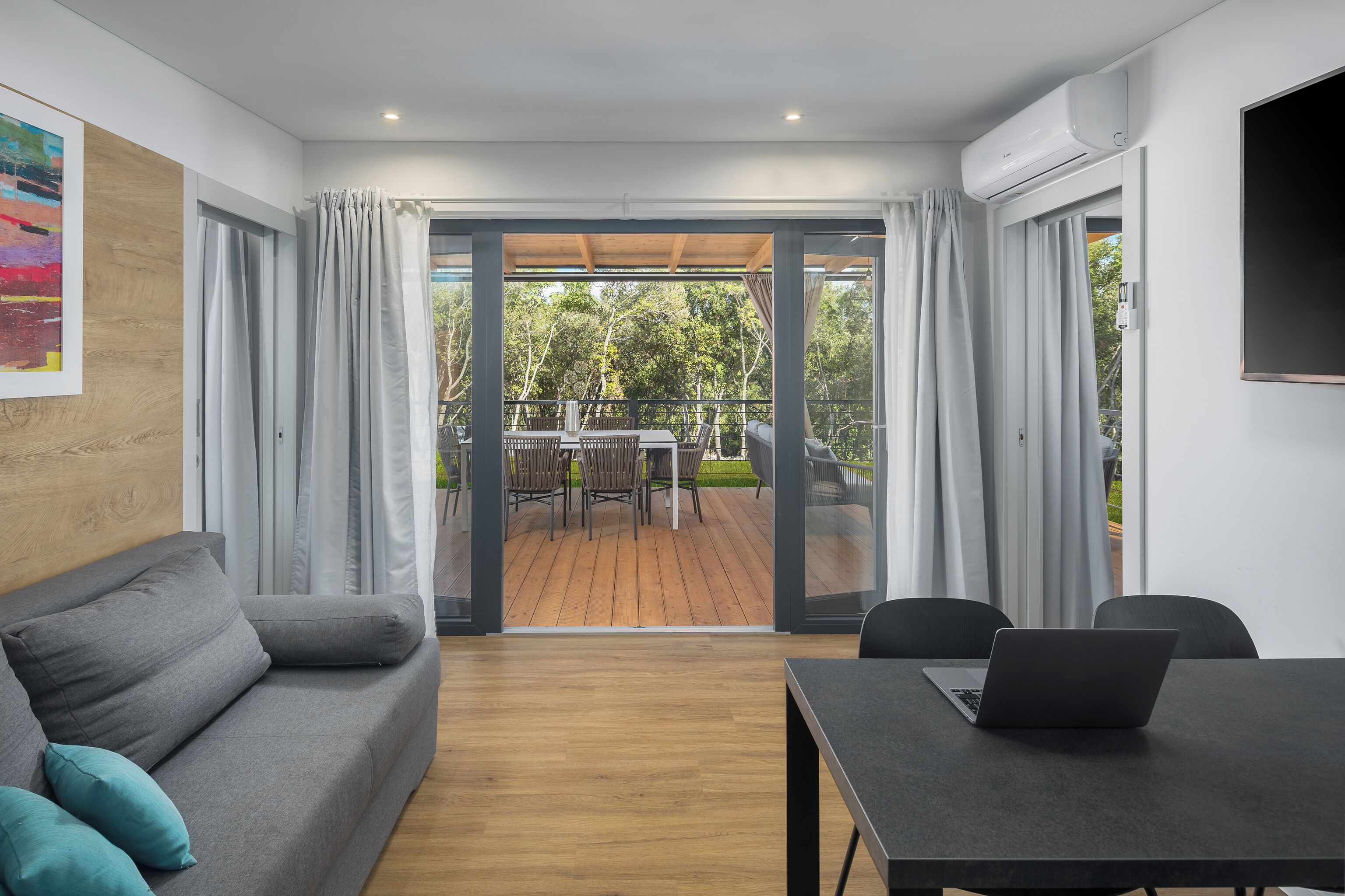 A modern room with a grey sofa, a table with a laptop, and a view of an outdoor deck with a table and chairs.
