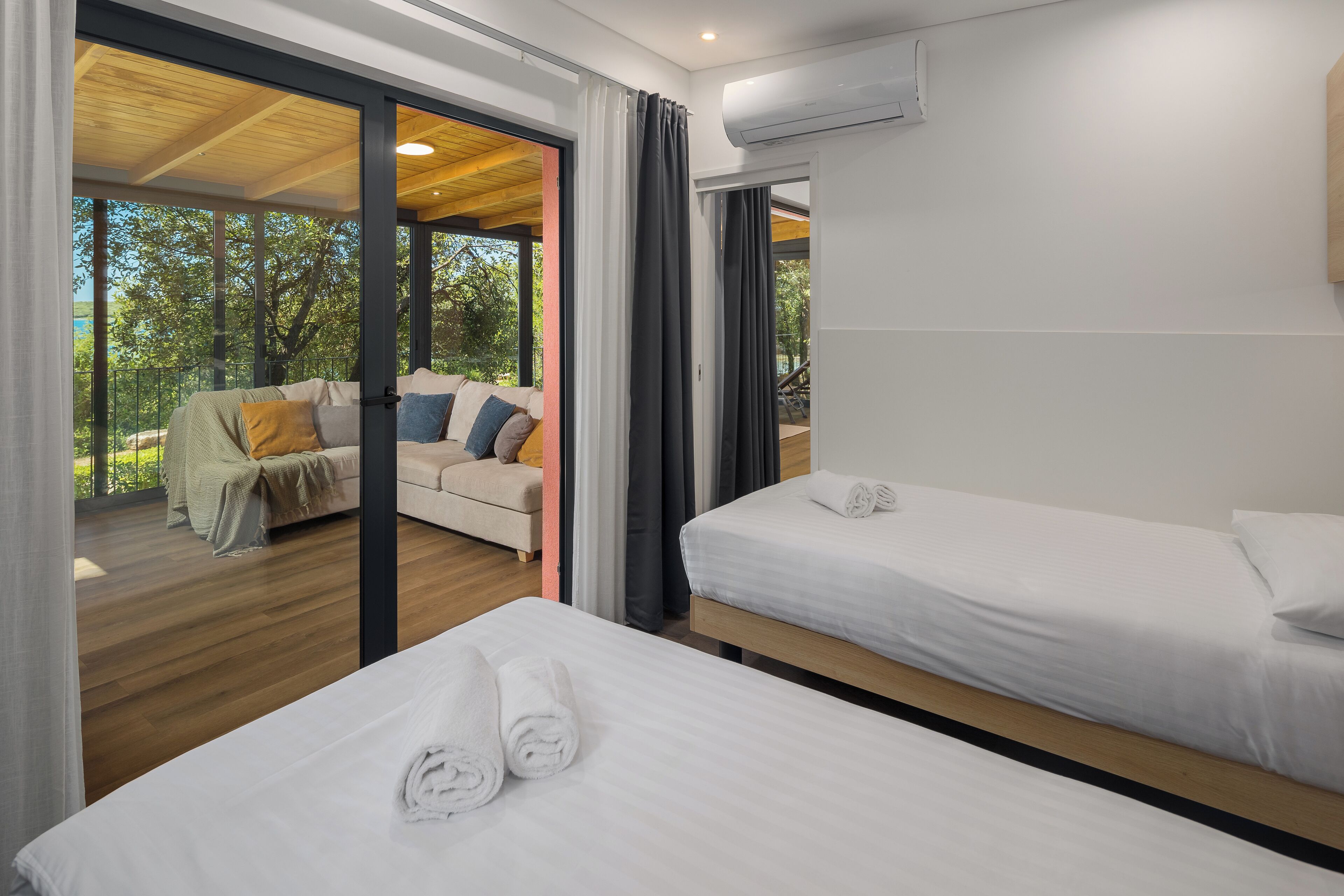 A modern hotel room with two beds, a balcony with a sofa, and a view of trees.