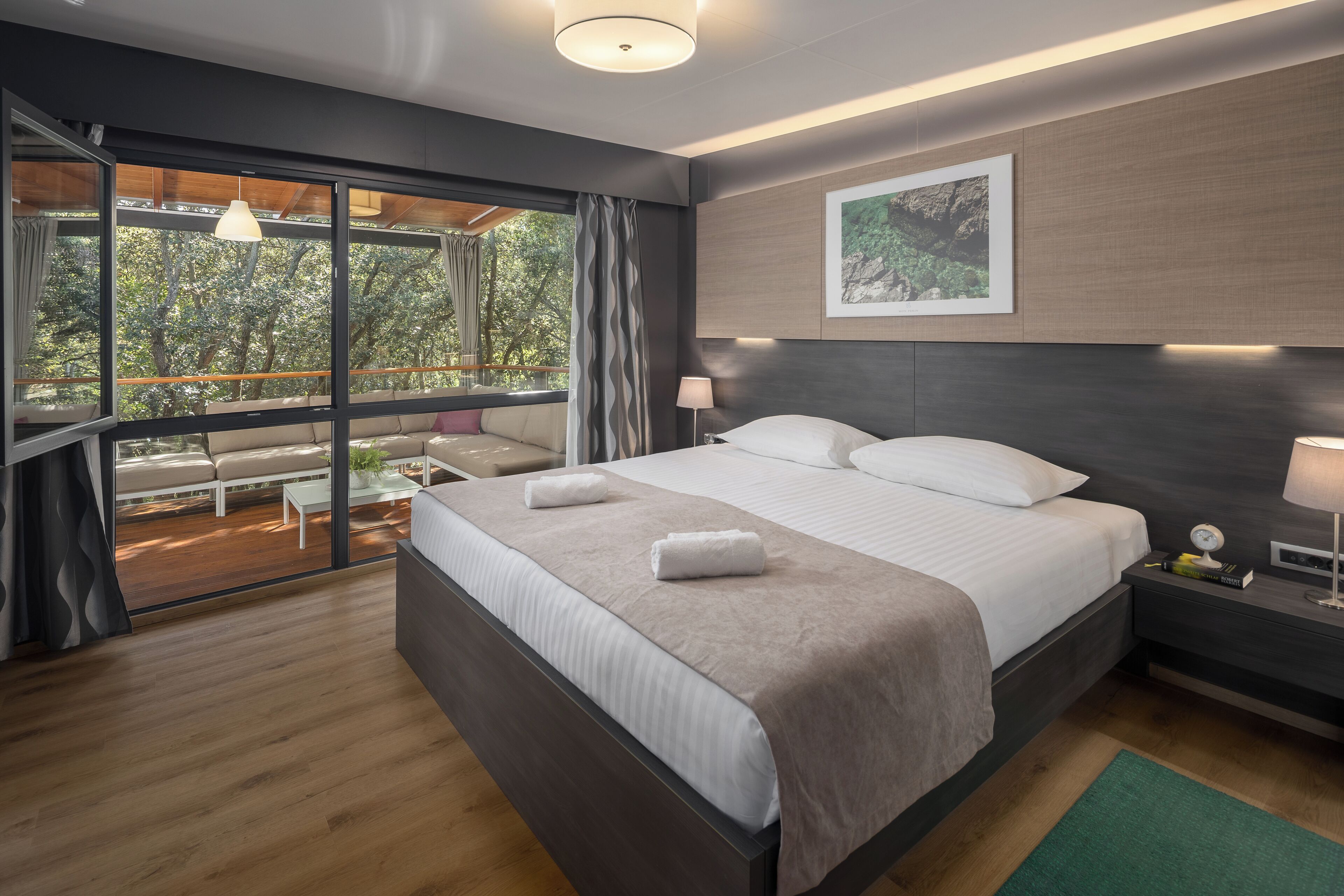 Un dormitorio moderno con una cama grande, una mesita de noche con lámpara, un cuadro en la pared y vistas al exterior a través de amplias ventanas.