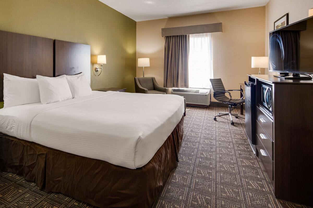 A hotel room with a large bed, two bedside lamps, a desk, a chair, a TV, and an air conditioning unit.