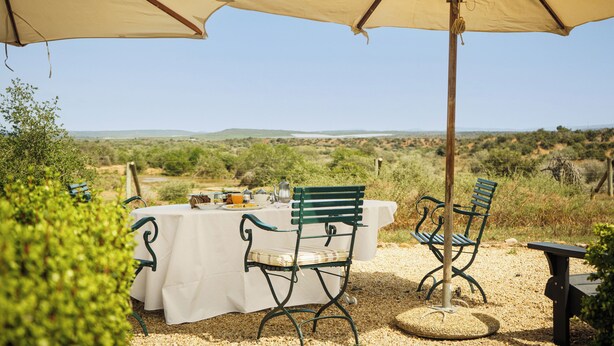 An outdoor dining area with a table set for two, surrounded by green metal chairs, under beige umbrellas, with a view of a hilly landscape. - RiverBend Lodge (Addo)