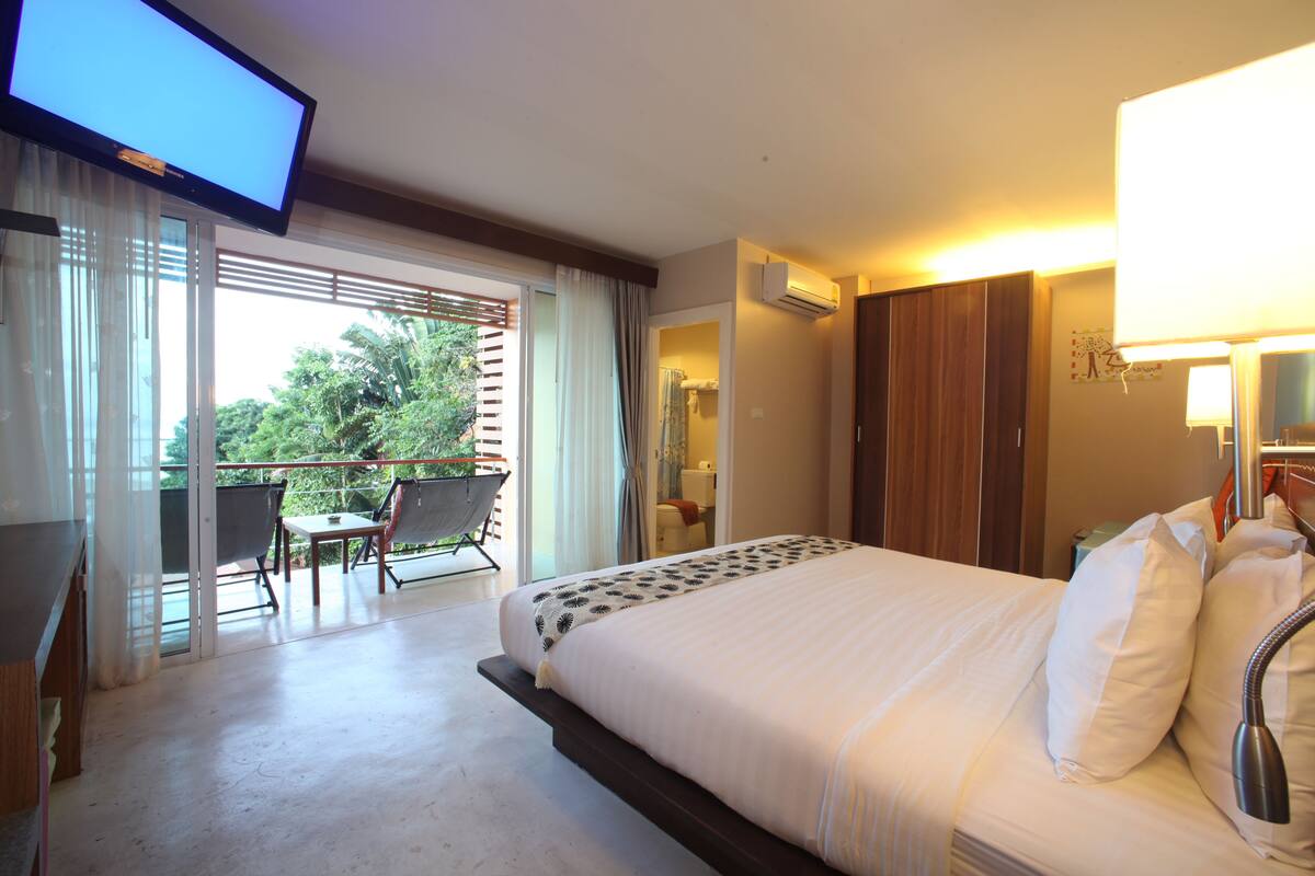 A hotel room with a bed, a TV, a balcony with a chair, and an air conditioning unit.