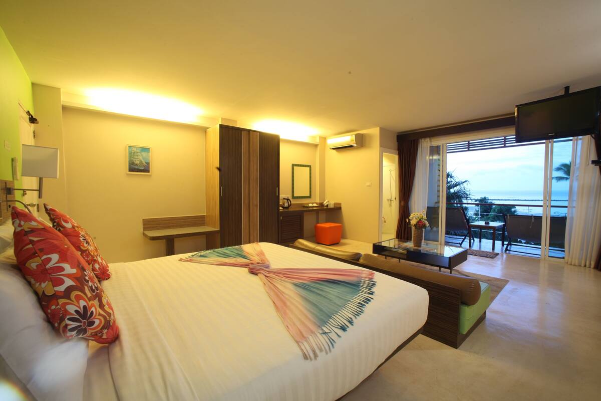 A hotel room with a bed, a desk, a chair, a small table, a TV, and a balcony with a view of the ocean.