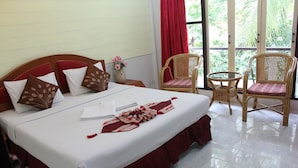 Una cama bien hecha con falda roja, ropa de cama blanca y cojines decorativos. Hay dos sillas de mimbre y una mesita redonda con un florero.