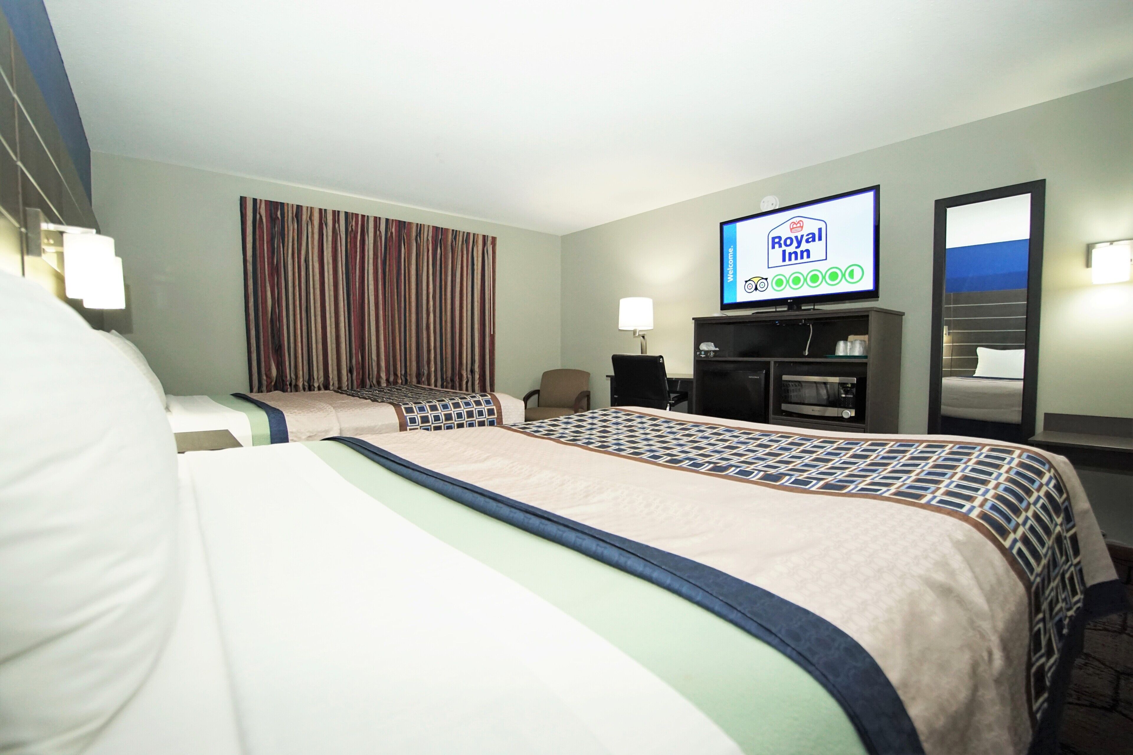 A hotel room with two beds, a television displaying the Royal Inn logo, a desk with a lamp, and a mirror.