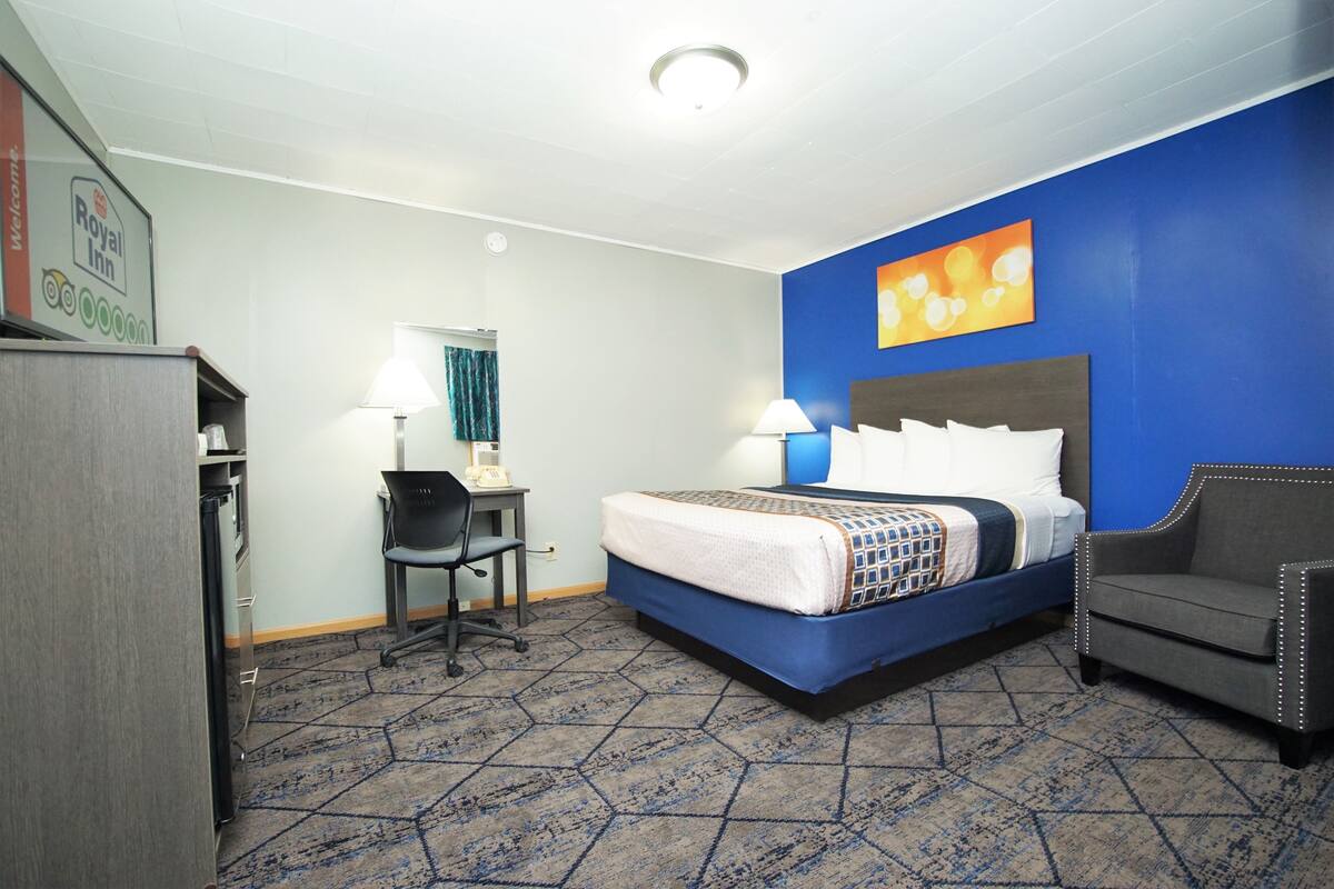 A hotel room with a bed, desk, chair, and a wall with a painting.