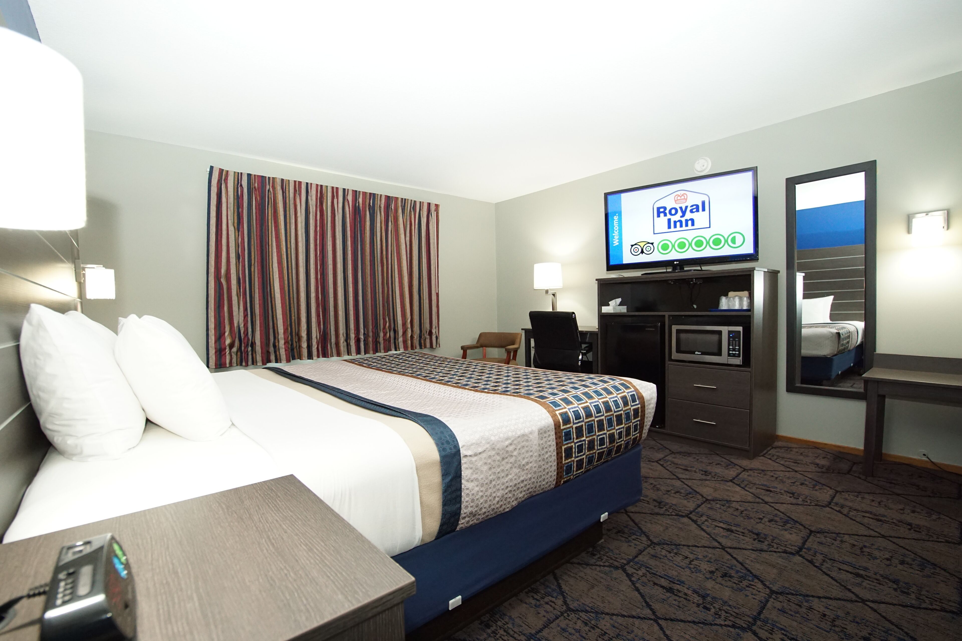 Una habitación de hotel con cama, escritorio, televisor mostrando el logotipo de Royal Inn, microondas, silla y cortina a rayas.