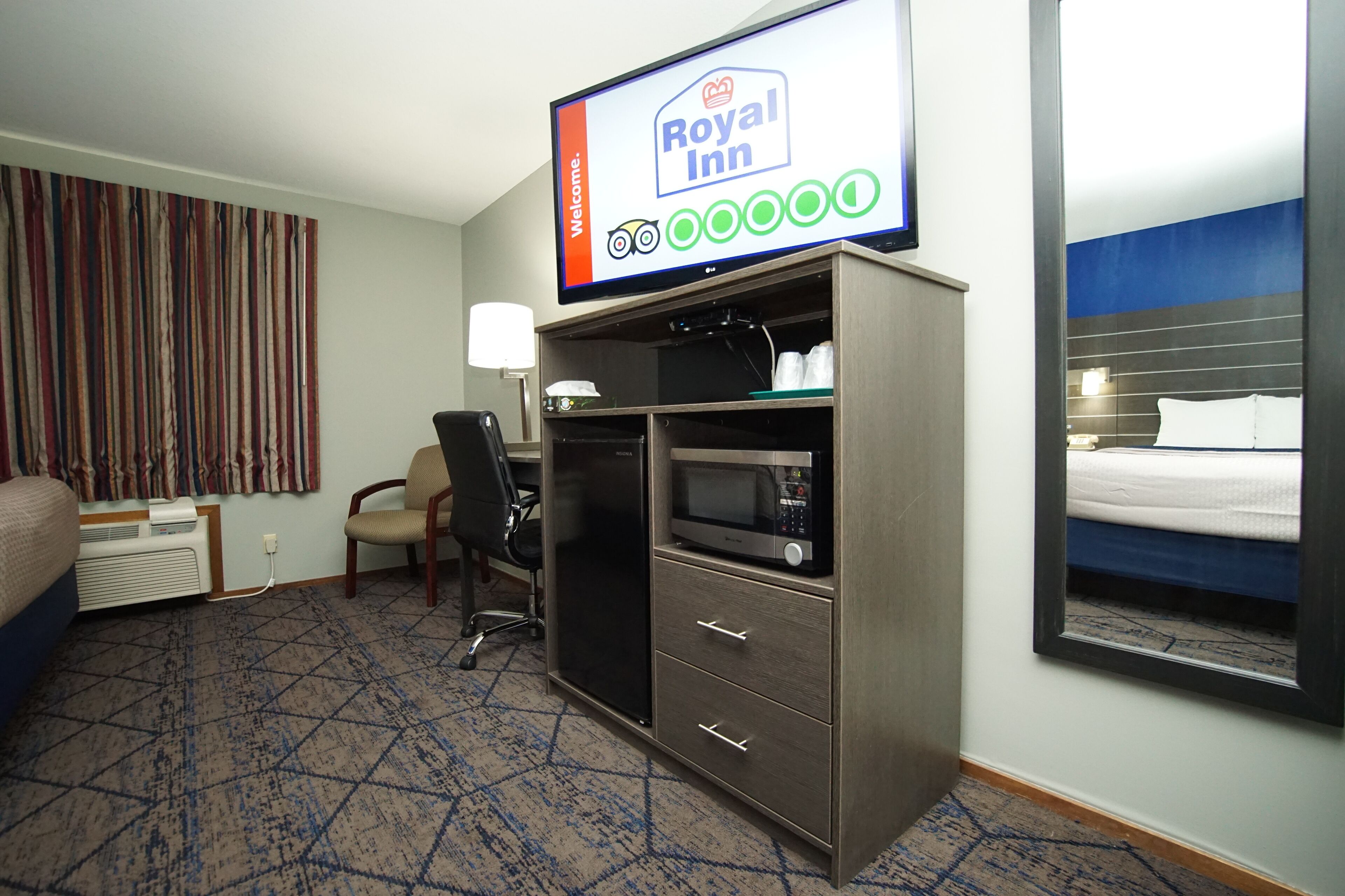 Une chambre d’hôtel équipée d’un lit, d’un bureau avec un ordinateur, d’un four à micro-ondes et d’un téléviseur affichant le logo de l’hôtel Royal Inn.