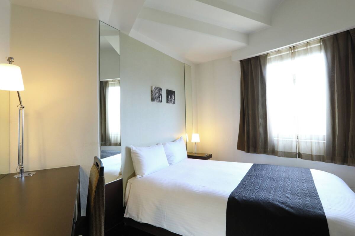 A hotel room with a large bed, a bedside table with a lamp, a mirror, and a window with curtains.