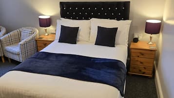 Un lit bien fait, avec une tête de lit sombre, deux tables de chevet avec des lampes, un fauteuil à carreaux et deux tableaux encadrés accrochés au mur.