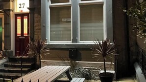 A building with a large illuminated sign reading "ACCOMMODATION" and a phone number. There are two potted plants in front of the entrance, and a small outdoor seating area with tables and chairs. - Q Accommodation (Blackpool)
