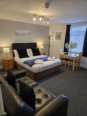 A hotel room with a bed, a desk with a chair, a coat rack, and a window with curtains. - Q Accommodation (Blackpool)