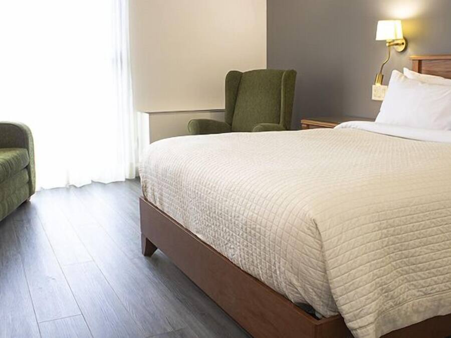 A hotel room with a large bed, a green armchair, a wooden nightstand, and a wall-mounted lamp.