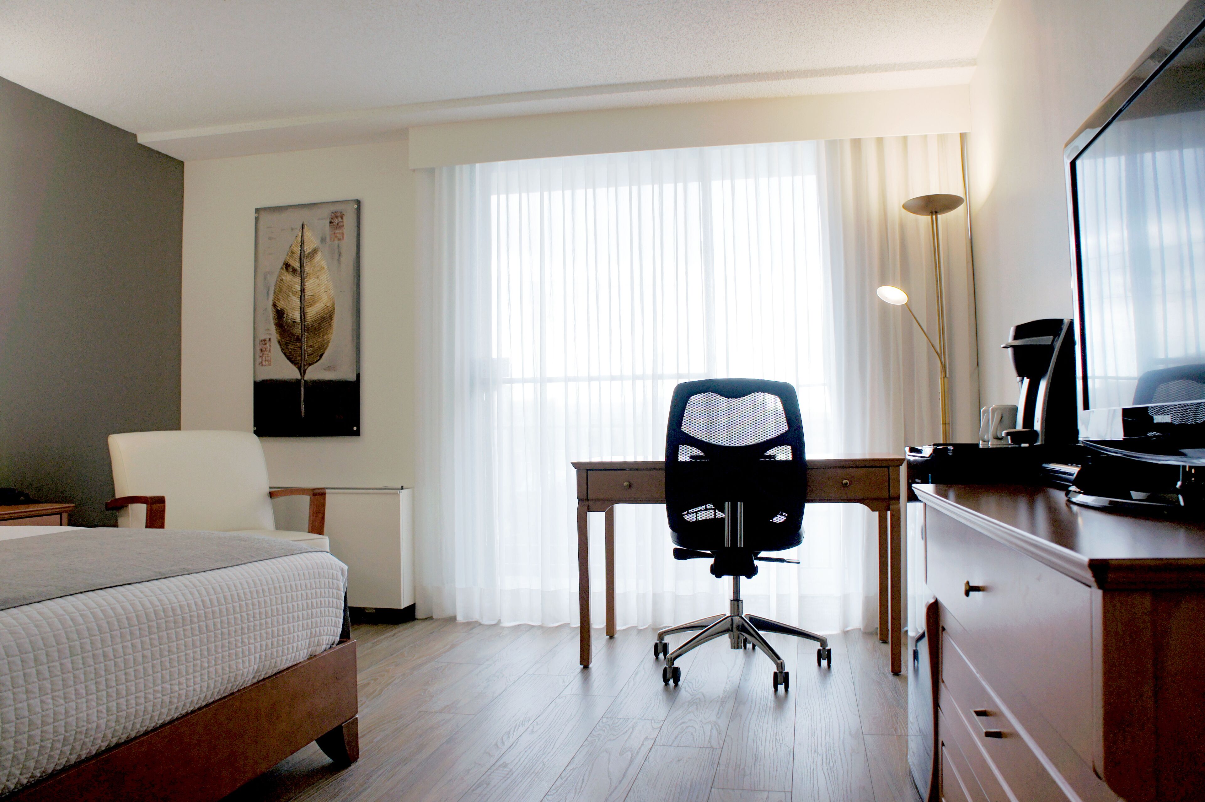 A hotel room with a bed, a desk with a chair, a lamp, and a painting on the wall.