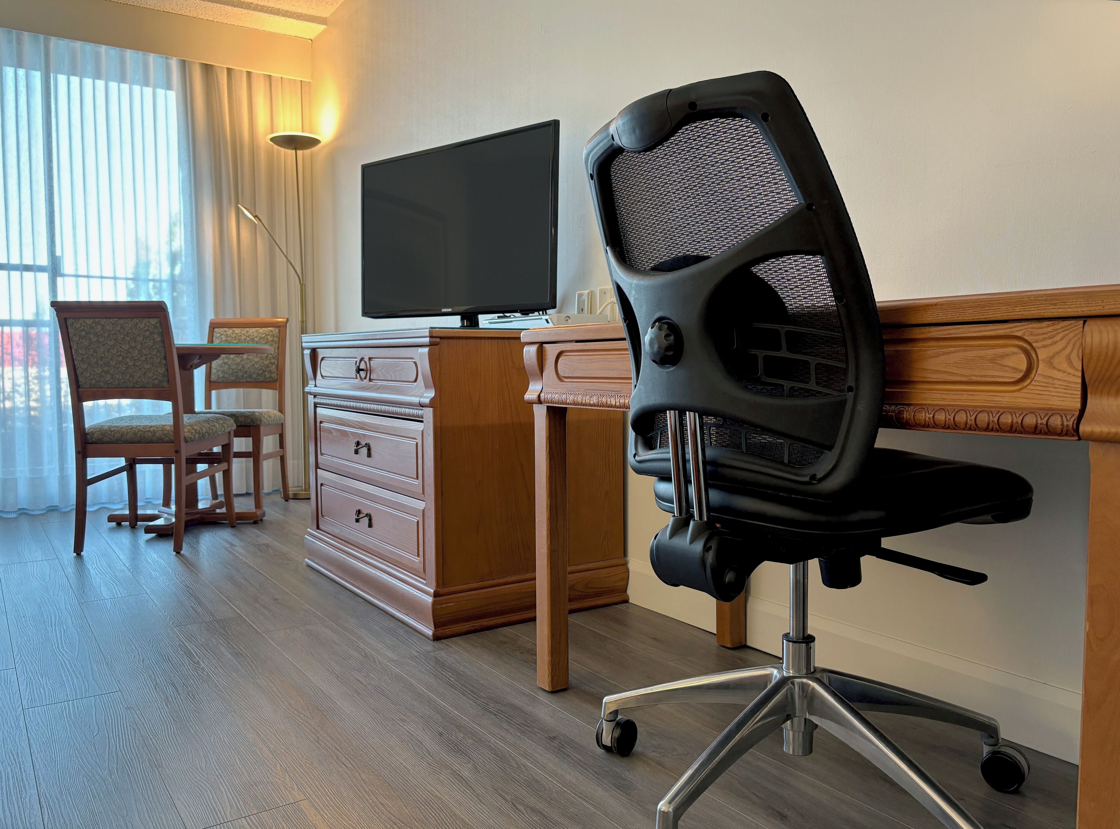 Une chambre d’hôtel avec un bureau en bois, une chaise de bureau noire, un téléviseur posé sur le bureau et une commode en bois avec des tiroirs.