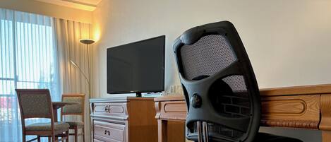 Habitación de hotel con un escritorio de madera, una silla de oficina negra, un televisor sobre el escritorio y un aparador de madera con cajones.