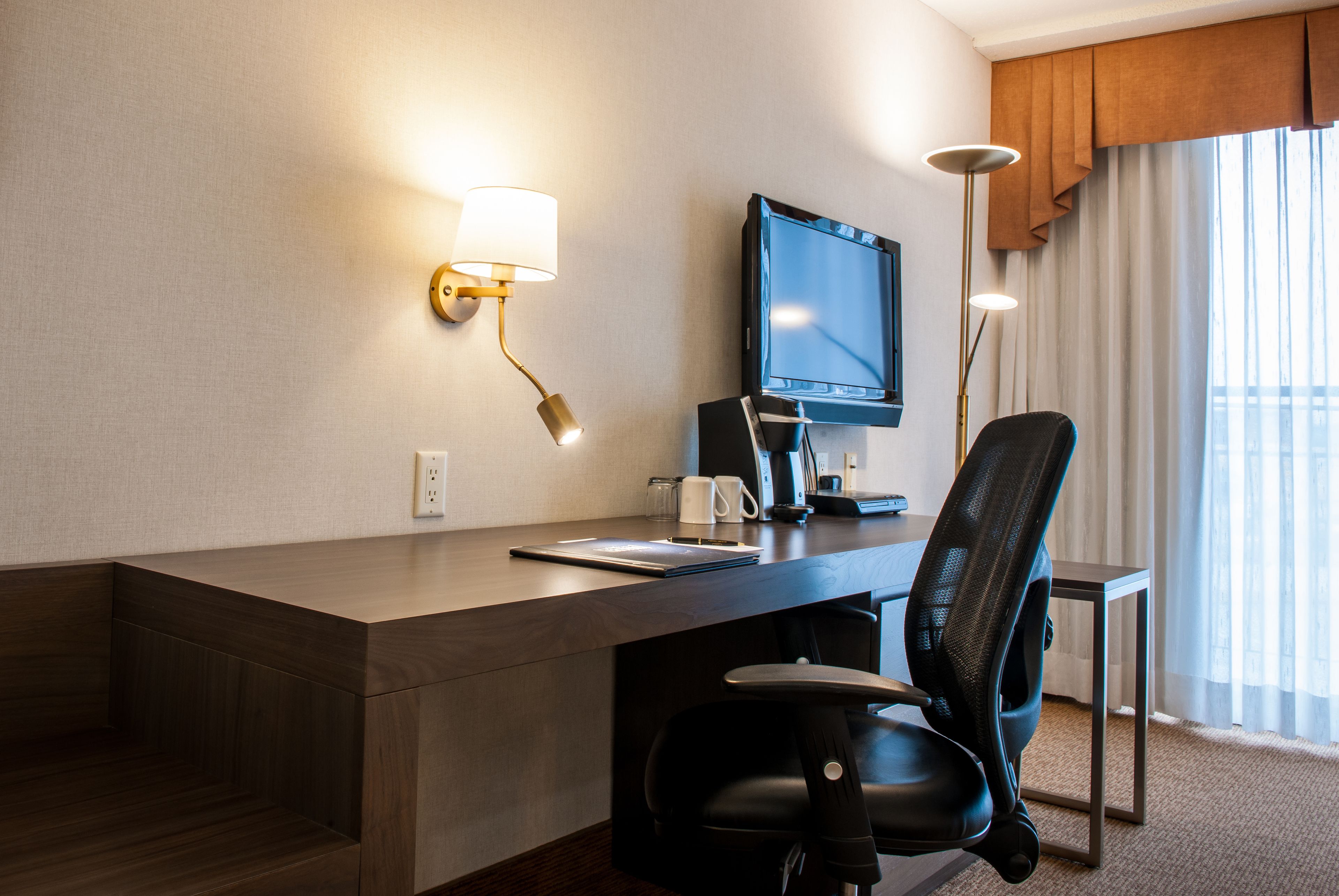 Una camera d'hotel con una scrivania in legno, una sedia da ufficio nera, una TV a schermo piatto, una lampada e una finestra con tende.