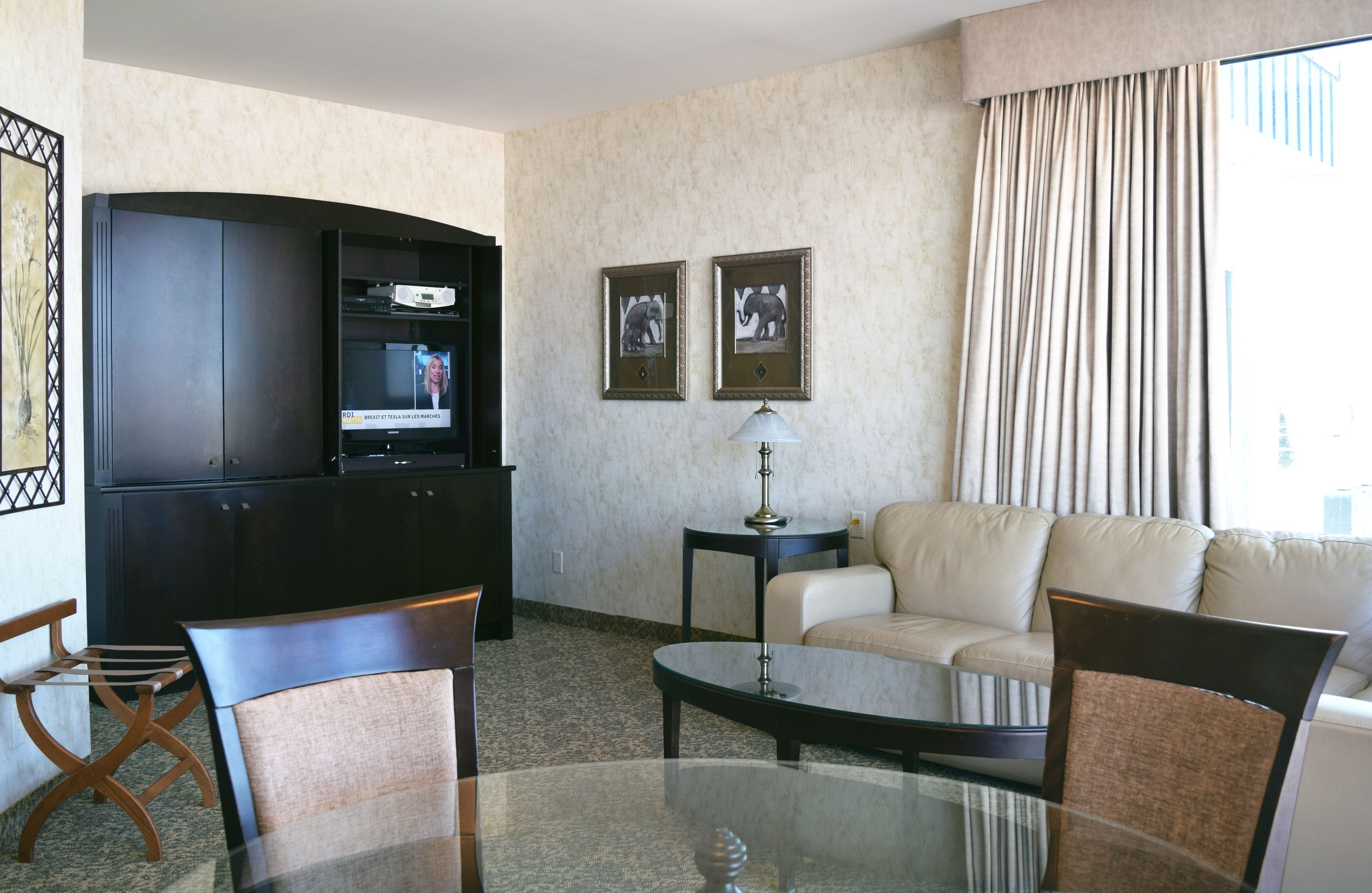 A hotel room with a glass coffee table, two chairs, a sofa, a TV, and framed pictures on the wall.