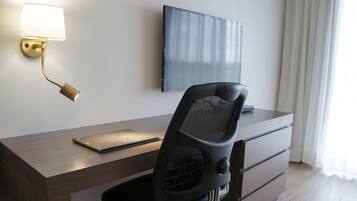 Uma mesa de escritório moderna com um laptop, uma cadeira de escritório preta, uma luminária fixada na parede e uma TV de tela plana na parede.