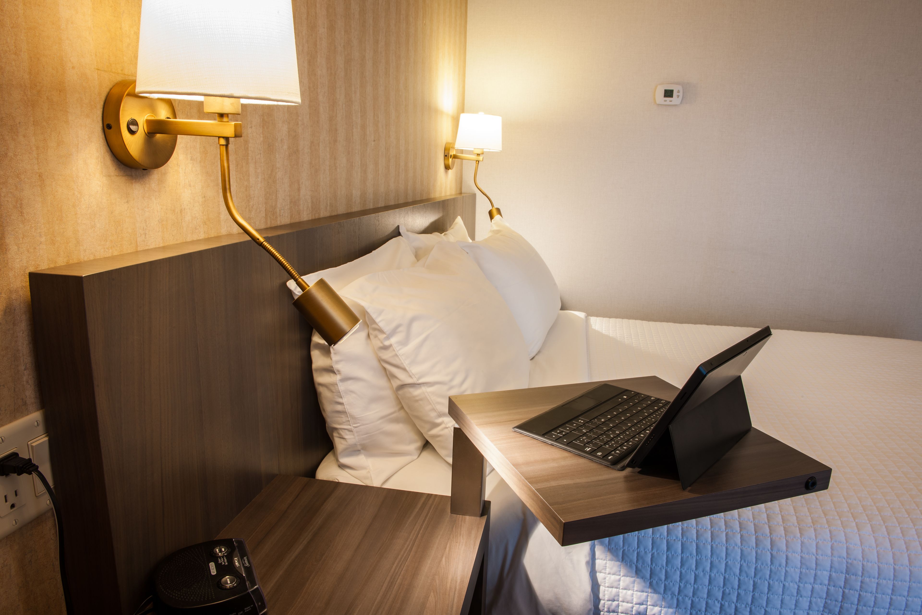 A hotel room with a bed, a bedside table with a laptop, a wall-mounted lamp, and a power outlet.