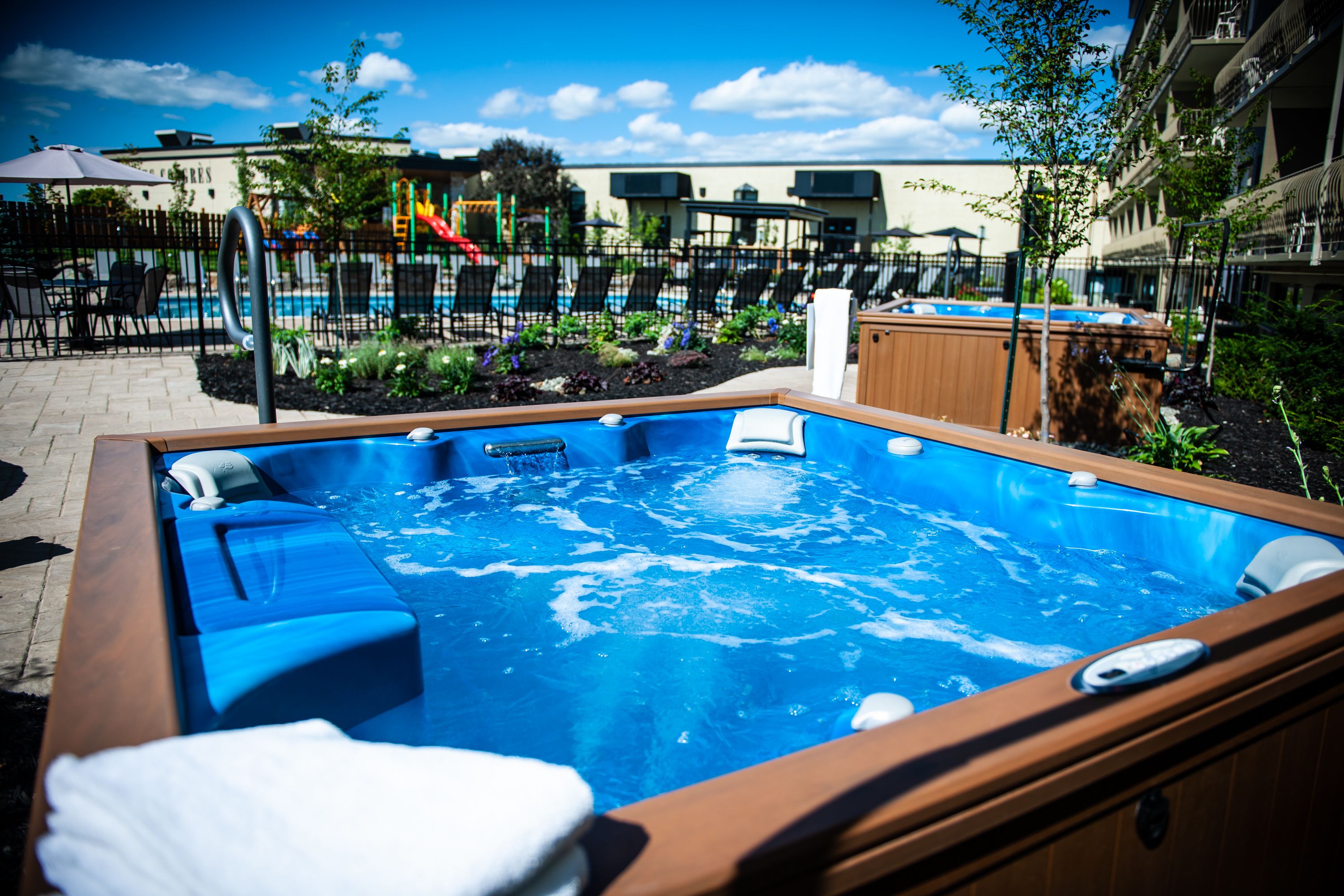 A hot tub with a wooden frame and blue cushions, surrounded by a fenced pool area with lounge chairs and a slide.