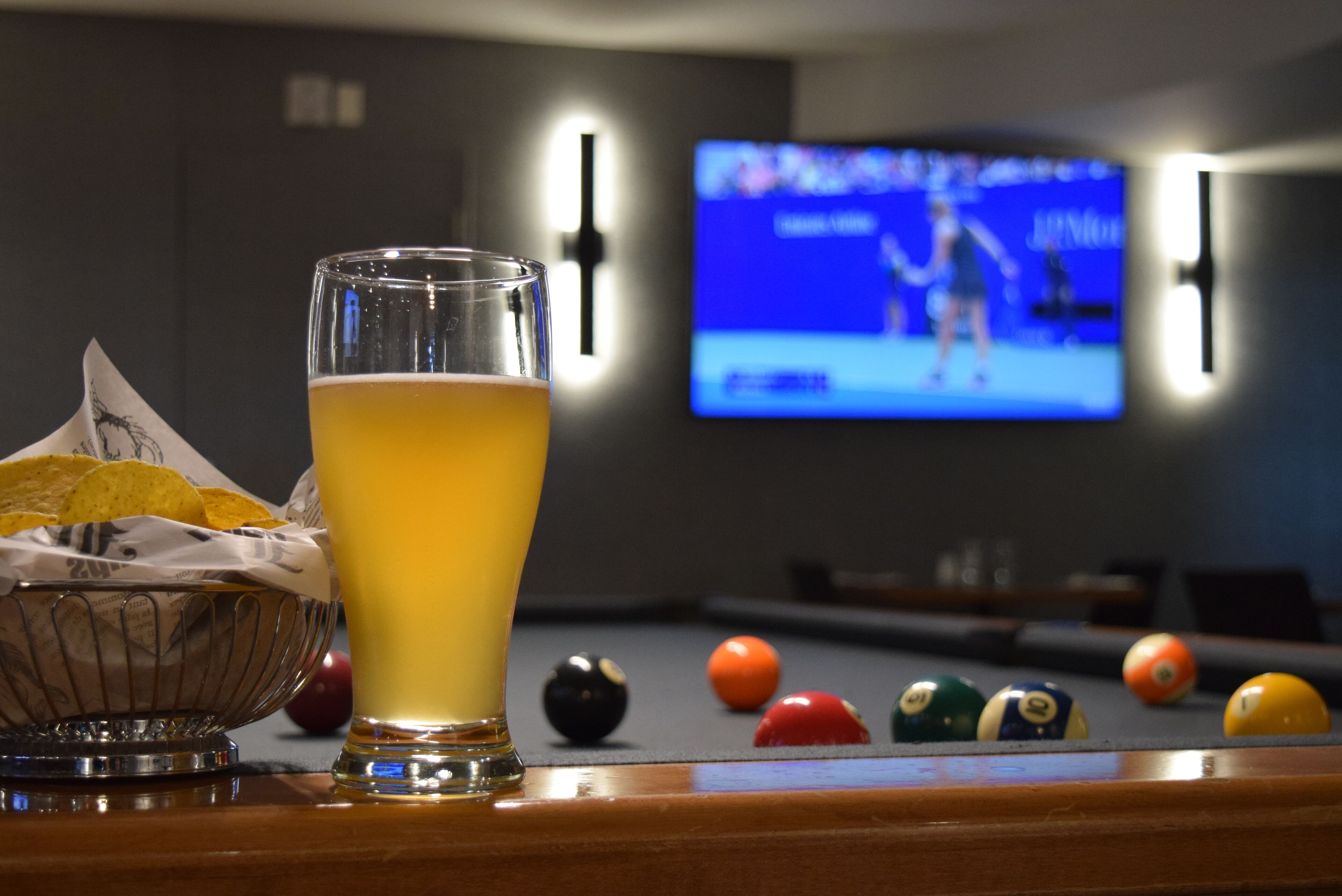 Un verre de bière, un bol de chips et une table de billard avec des billes, dans une pièce faiblement éclairée où un écran de télévision diffuse un événement sportif.