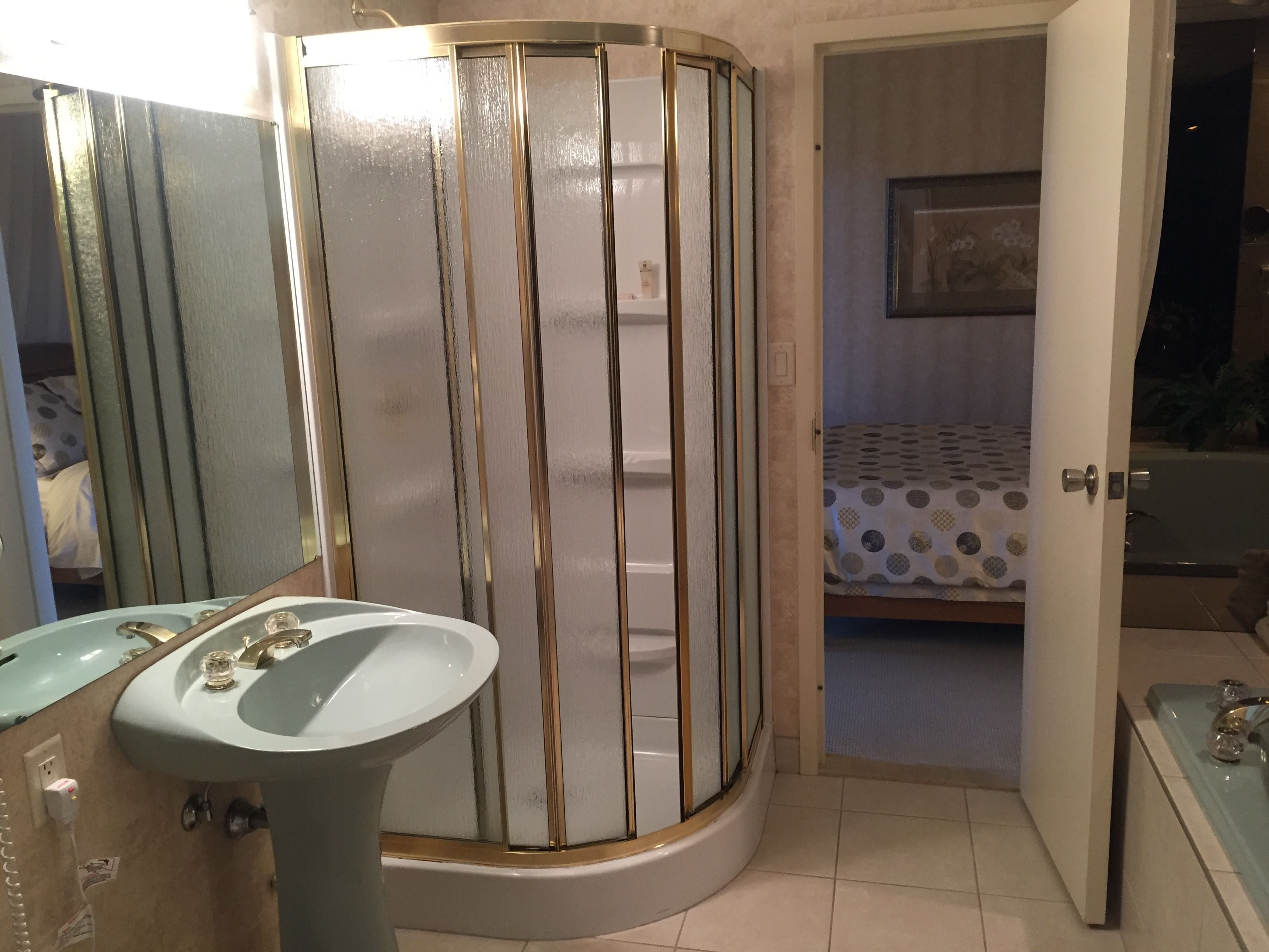 Un baño con lavabo, espejo, cabina de ducha y una cama visible a través de una puerta abierta.