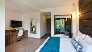 A modern hotel room with a large bed, a desk with a TV, a mirror, and a view of the outdoors.