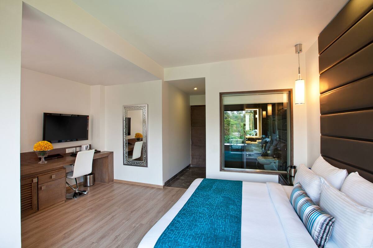 Habitación de hotel moderna con una cama grande, un escritorio con televisor, un espejo y vistas al exterior.