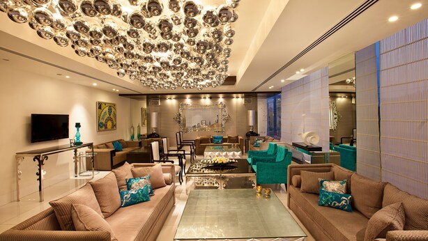 A modern living room with a glass coffee table, two sofas with patterned cushions, a large chandelier, and a television on the wall.