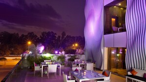An outdoor dining area with tables and chairs, a modern building with wavy architectural features, and a night sky.