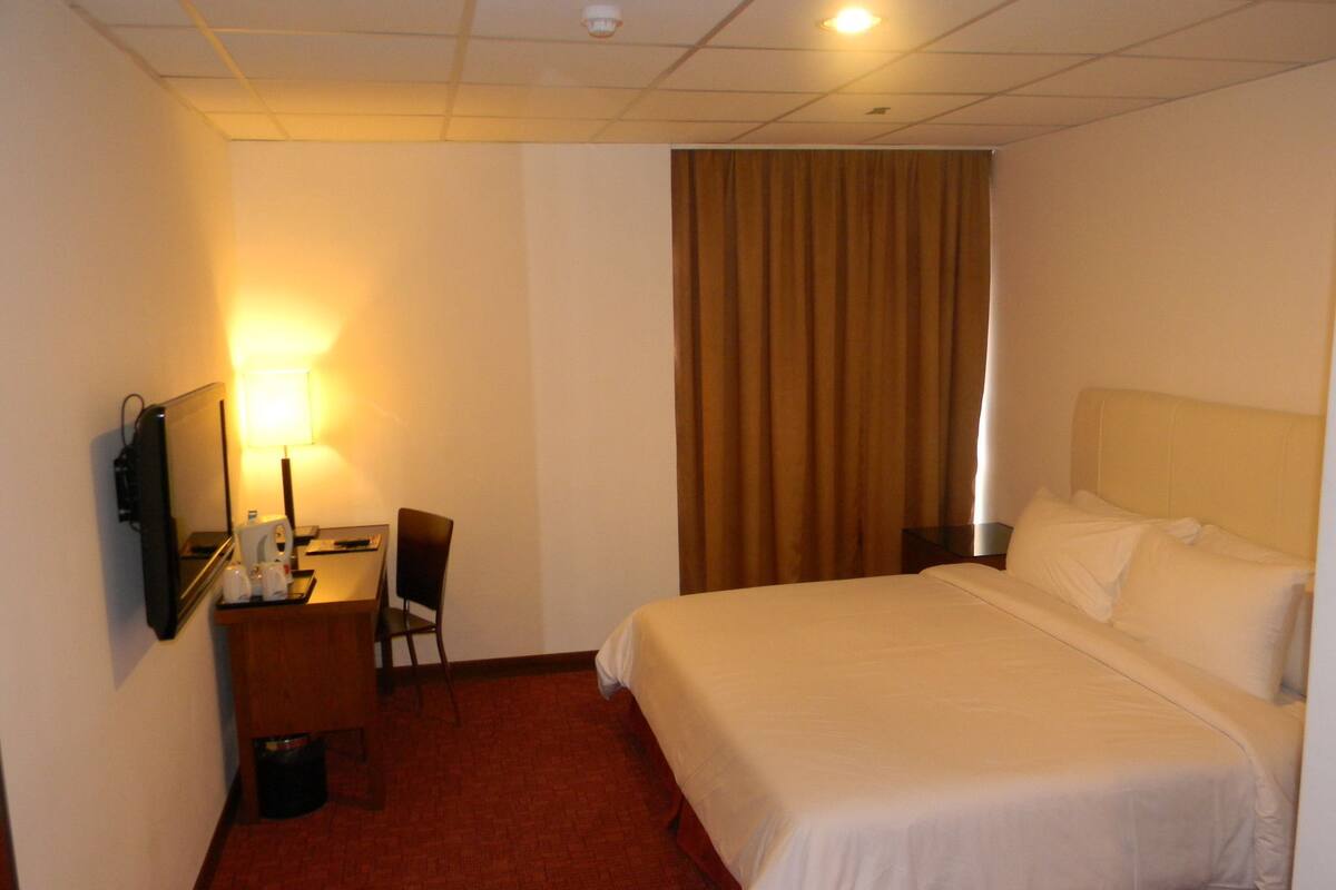 A hotel room with a bed, a desk with a chair, a television, and a window with curtains.