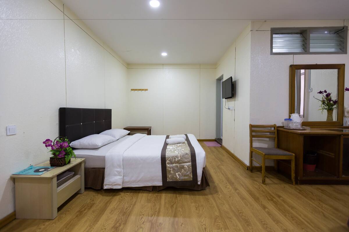 A bedroom with a bed, bedside table, chair, desk, mirror, and a TV mounted on the wall.