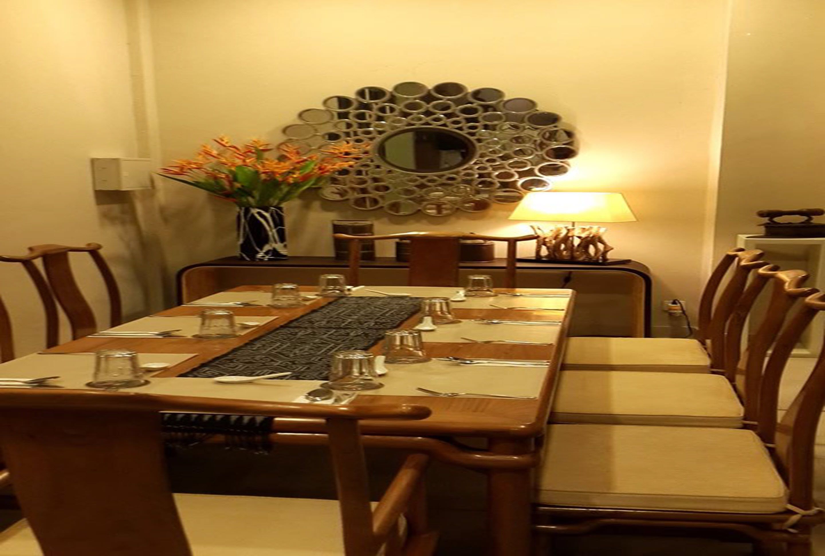 A dining area with a long table set for six, surrounded by wooden chairs with cushioned seats. A decorative mirror and a vase with flowers are on the wall behind the table.