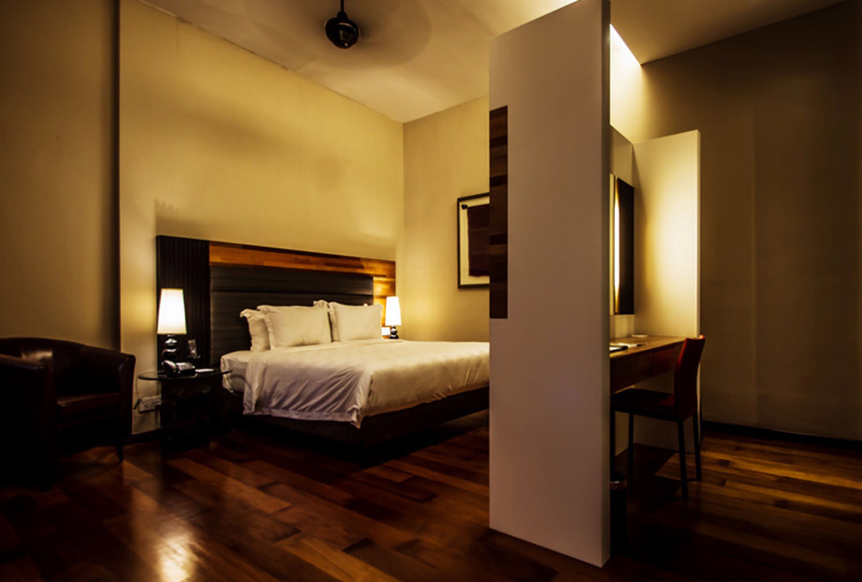 A hotel room with a wooden floor, a bed with white linens, a desk with a chair, and a nightstand with a lamp.