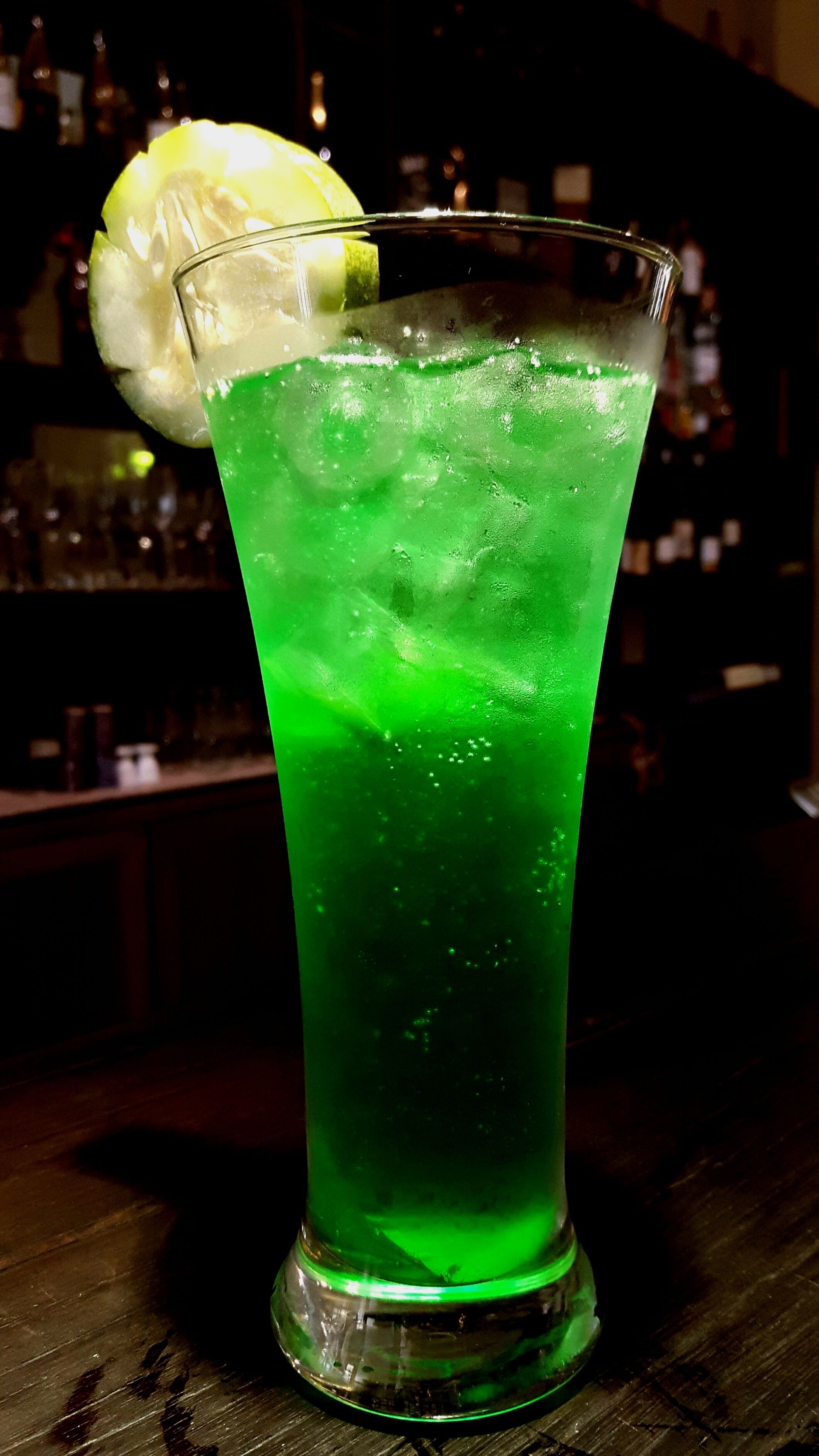 A tall glass with a green beverage, ice cubes, and a lemon slice garnish on a bar counter.