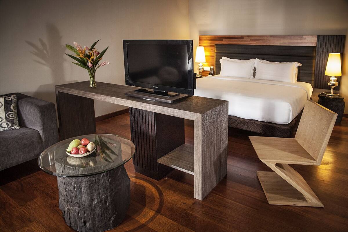 A hotel room with a bed, a TV on a wooden table, a sofa, a small round table with fruit, and a lamp.