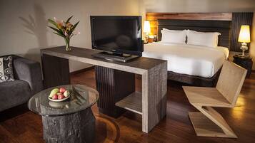 Habitación de hotel con cama, televisor sobre una mesa de madera, sofá, una mesita redonda con fruta y una lámpara.