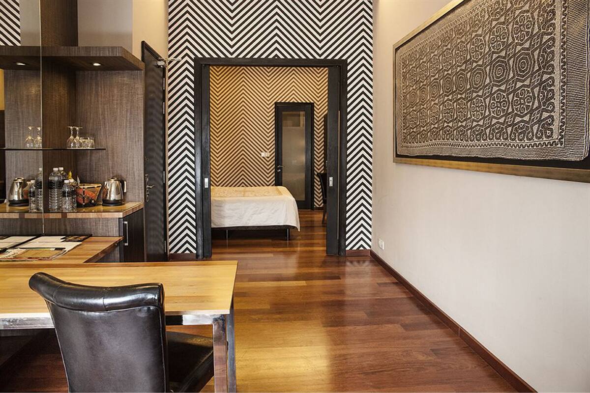 A modern hotel room with a wooden floor, a bed with a white bedspread, a dining area with a table and chairs, and a kitchen area with a counter and various items.