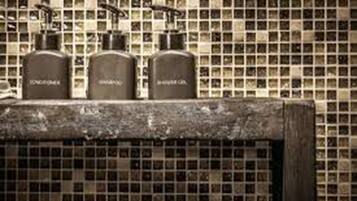 A bathroom with a tiled backsplash and three bottles labeled "CONDOMINIUM," "SHAMPOO," and "SHOWER GEL" on a shelf.