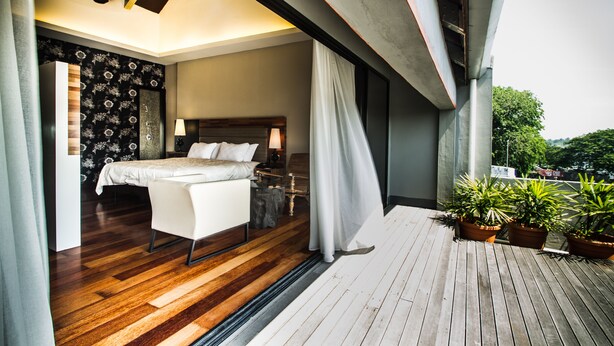Un dormitorio moderno con suelo de madera, una cama grande, una mesita de noche y una ventana con cortinas. Hay plantas en macetas en el balcón.