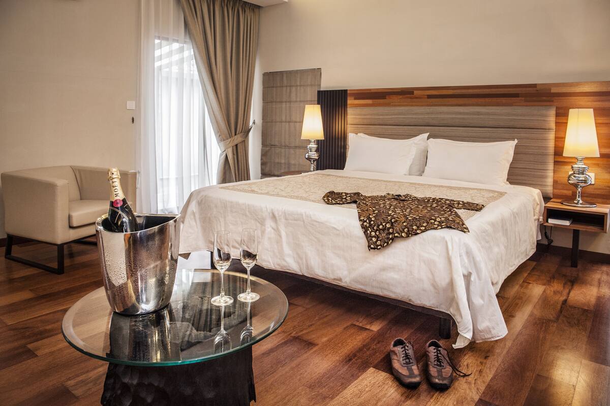 A hotel room with a large bed, two bedside lamps, a glass table with a bottle and glasses, a chair, and a pair of shoes on the floor.